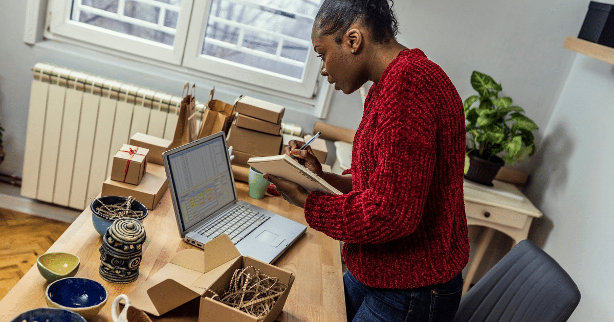 Woman in a red sweater taking notes while managing business tasks at a desk with a laptop and packaging materials.