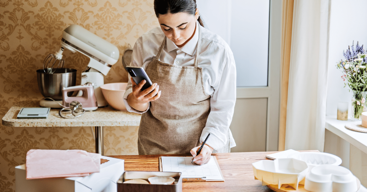 Woman in an apron using a phone while writing notes in a kitchen, preparing for a baking project.