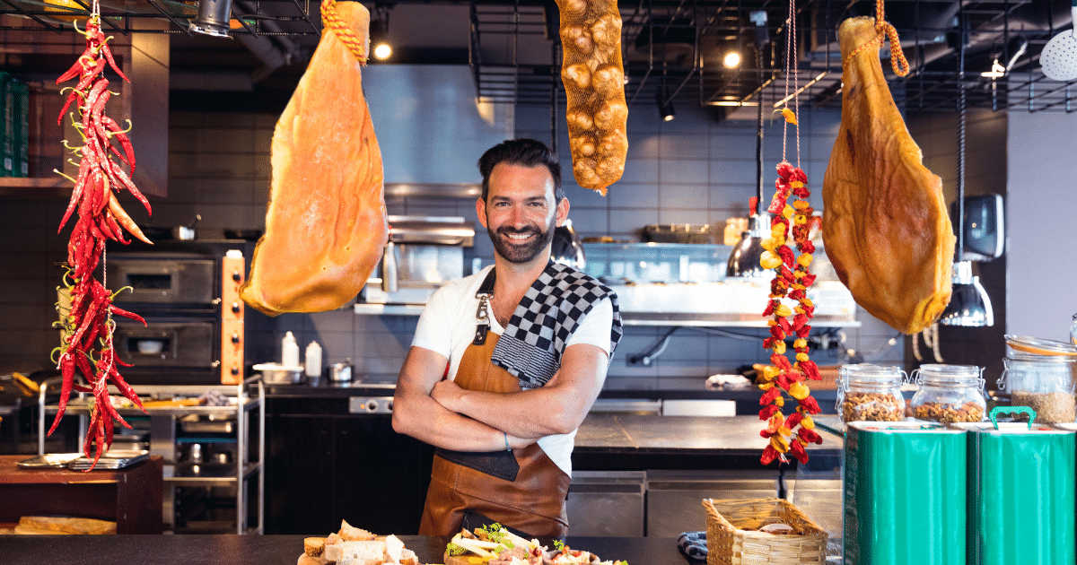 Chef in a restaurant kitchen with hanging meats and spices, showcasing effective business budget strategies for culinary success.