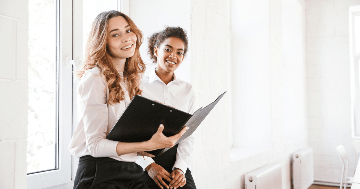Two businesswomen smiling and discussing strategic holiday budgeting in a bright office setting.
