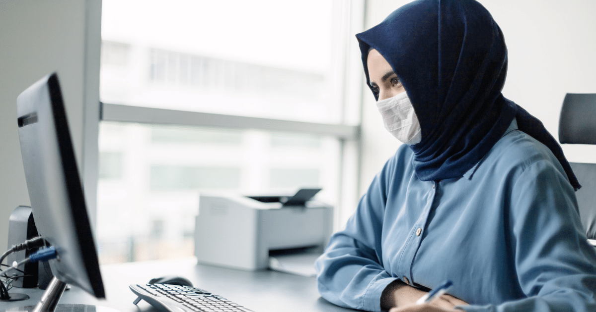 Woman in a blue shirt and mask working at a computer, representing small business growth and strategic planning.