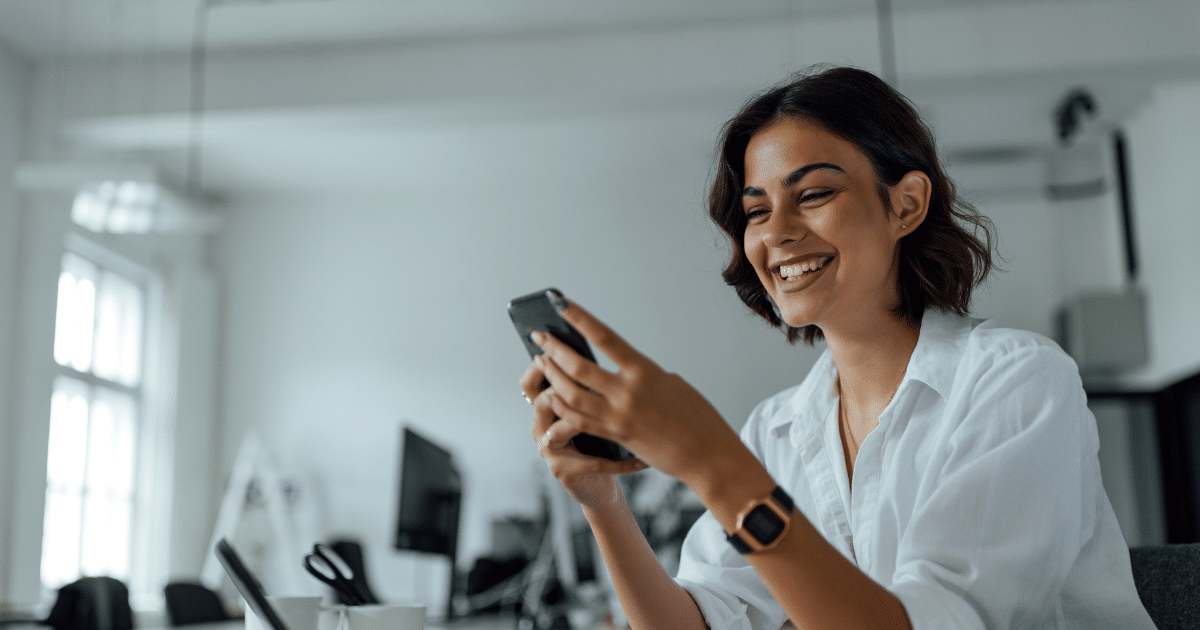Smiling woman in a white shirt engaging with her phone, representing successful dealmaking and sales strategies.