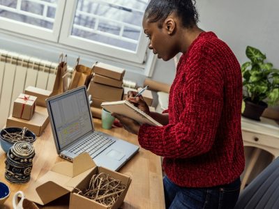 Woman in a red sweater taking notes while managing business tasks at a desk with a laptop and packaging materials.