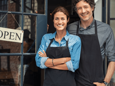 Smiling grocery business owners standing by an open sign, representing bad credit business loans for growth and cash flow.