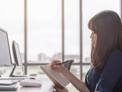 Businesswoman taking notes at a desk with computers, emphasizing profitable business habits and financial growth strategies.